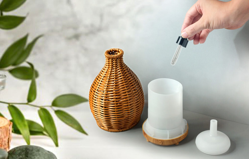 Na obrazie widoczny jest aromatyzer w kształcie wazonu z wikliny w kolorze brązowym, obok którego znajduje się biały pojemnik i ręka trzymająca pipetę. W tle widać zielone liście na rozmytym tle.