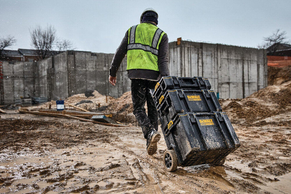 Skrzynka narzędziowa DEWALT DS450 plac budowy błoto osoba ciągnie za sobą trzy skrzynki w tle betonowe konstrukcje skuteczny system uszczelnień ochrona przed wnikaniem pyłu wilgoci i innych zanieczyszczeń wydłuża żywotność sprzętu ogranicza ryzyko korozji uszkodzeń i konieczności częstych napraw zawartość pozostaje sucha i czysta