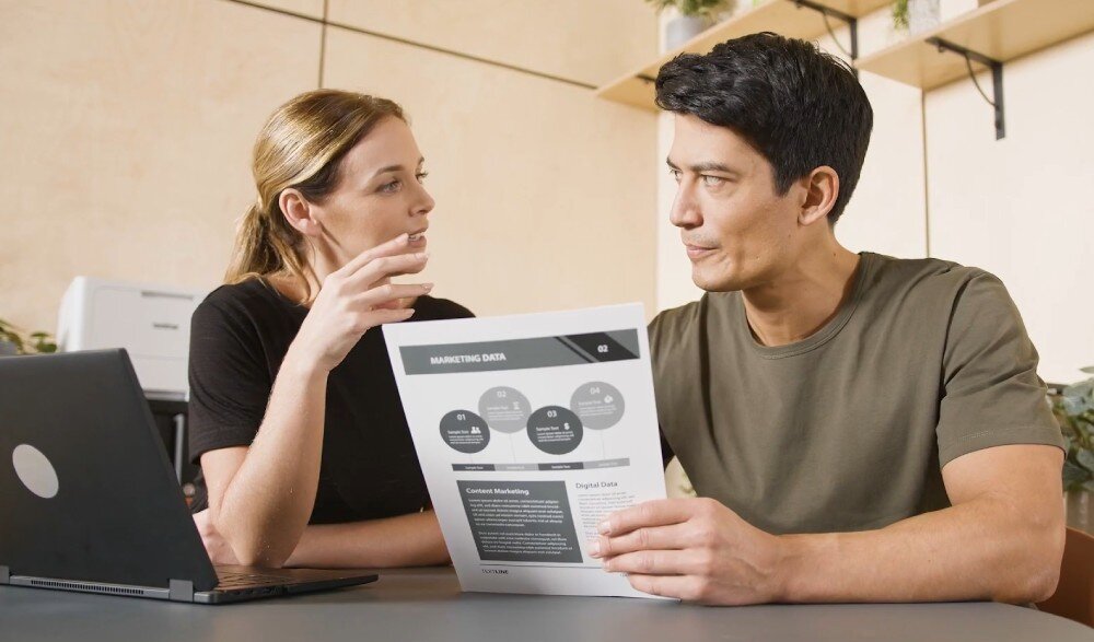 Drukarka BROTHER HL-L5210DW kobieta i mężczyzna rozmawiający i siedzący przy stoliku, na którym leży laptop, mężczyzna trzyma karki z czarno-białym wydrukiem zabezpieczenia głośność