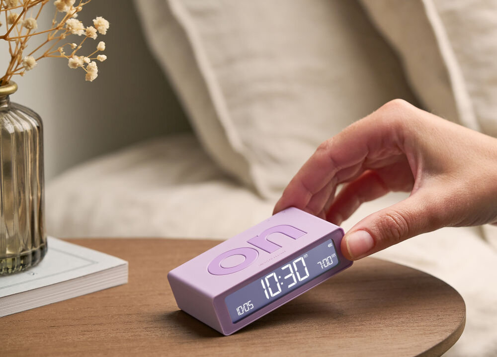 Budzik LEXON Flip Mini liliowy, ergonomia, ON, OFF, obrót, Ręka przechyla mały budzik Lexon Flip Mini w kolorze grafitowym, stół, książka, neutralne tło.