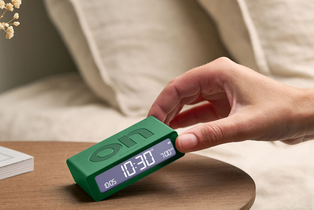 Budzik LEXON Flip Mini zielony, ergonomia, ON, OFF, obrót, Ręka przechyla mały budzik Lexon Flip Mini w kolorze grafitowym, stół, książka, neutralne tło.