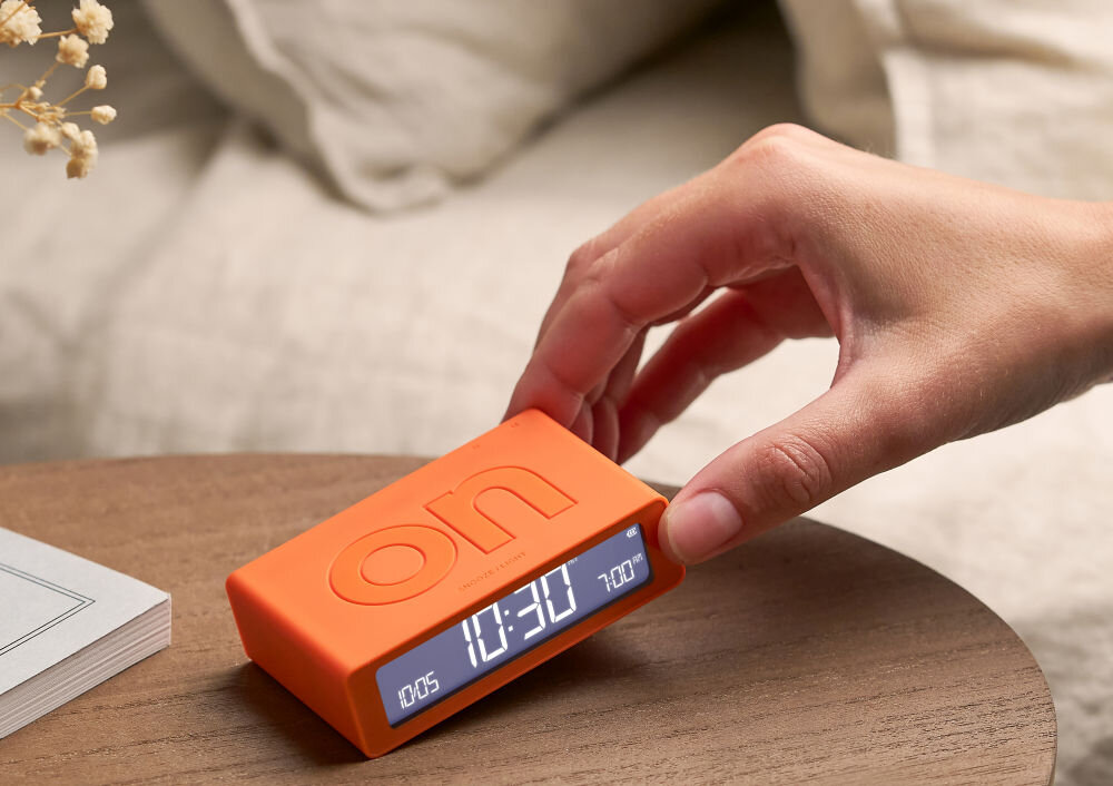 Budzik LEXON Flip Mini pomarańczowy, ergonomia, ON, OFF, obrót, Ręka przechyla mały budzik Lexon Flip Mini w kolorze grafitowym, stół, książka, neutralne tło.