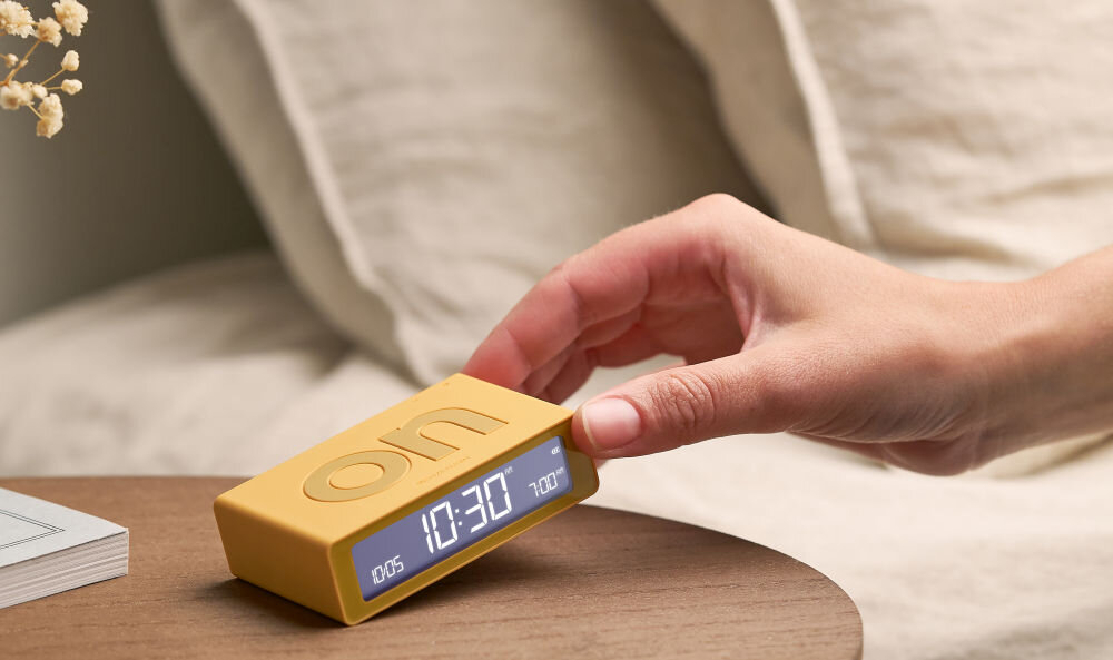 Budzik LEXON Flip Mini żółty, ergonomia, ON, OFF, obrót, Ręka przechyla mały budzik Lexon Flip Mini w kolorze grafitowym, stół, książka, neutralne tło.