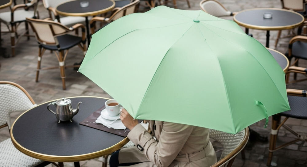 Parasolka LEXON Mini Hook LU21LL miętowy, ergonomia, forma, deszcz, pogoda, kobieta z parasolką w dłoni, kobieta trzyma parasolkę w dłoni siedząc przy stoliku na zewnątrz placu