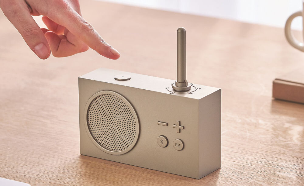 Radioodtwarzacz LEXON Tykho 3 złoty, design, matowe wykończenie, styl, minimalizm, Palec dotyka przycisku na radiu Lexon Tykho 3, panel z głośnikiem, filiżanka w tle.