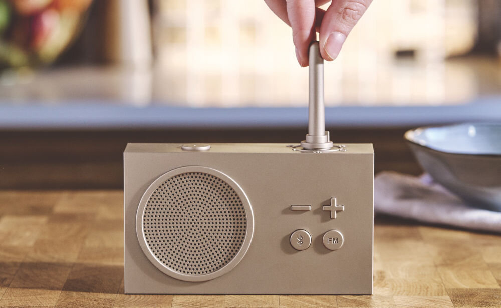 Radioodtwarzacz LEXON Tykho 3 złoty, radio, Bluetooth, głośnik, funkcjonalność, Ręka obraca gumową antenę radia Lexon Tykho 3, widok z przodu, blat kuchenny, przyciski sterujące.