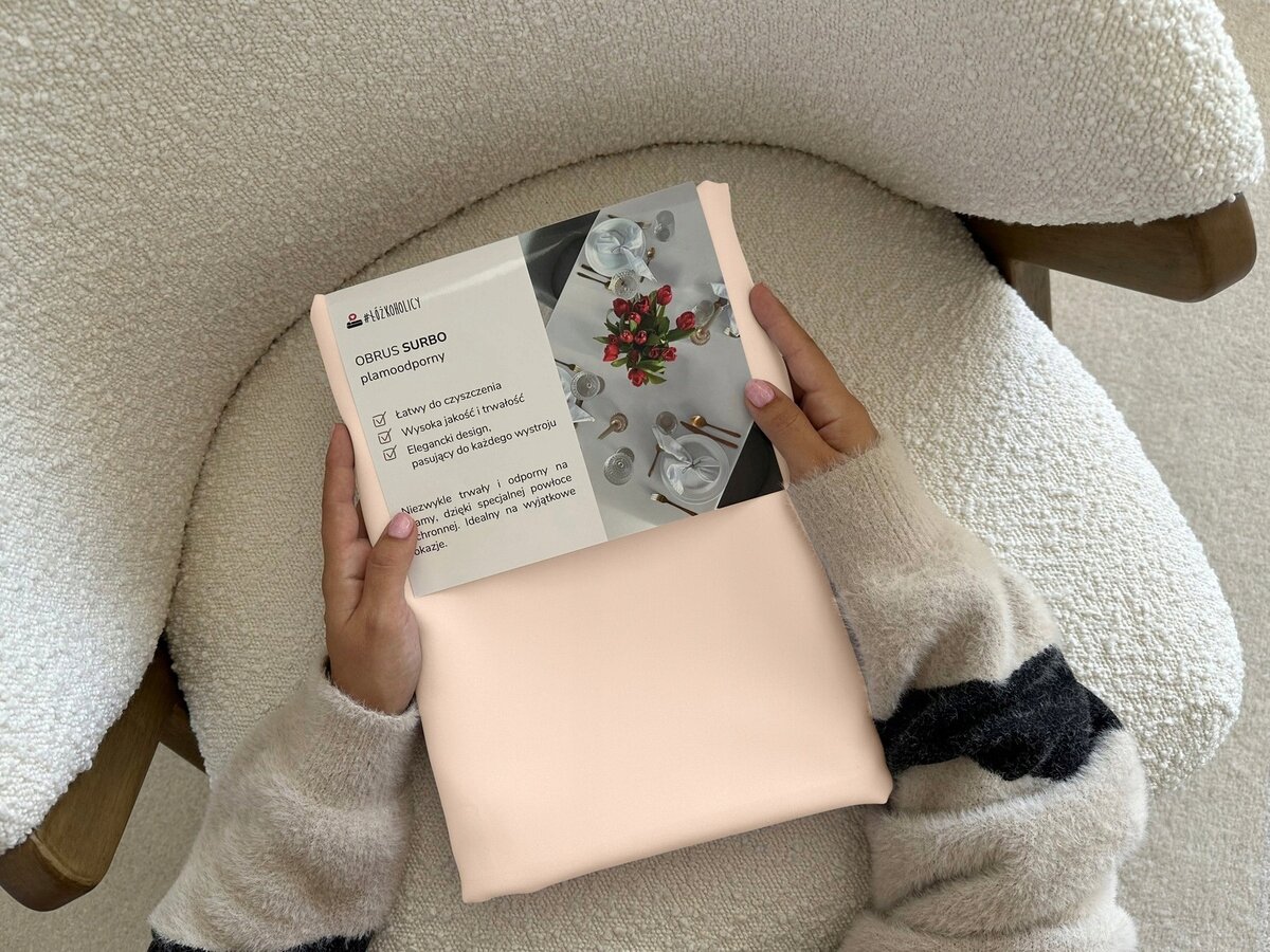 Osoba trzyma opakowanie z napisem \'OBRUS SURBO plamoodporny\', na którym widnieje zdjęcie nakrytego stołu z kwiatami. Opakowanie zawiera informacje o łatwości czyszczenia, wysokiej jakości i dopasowaniu do każdego wystroju.