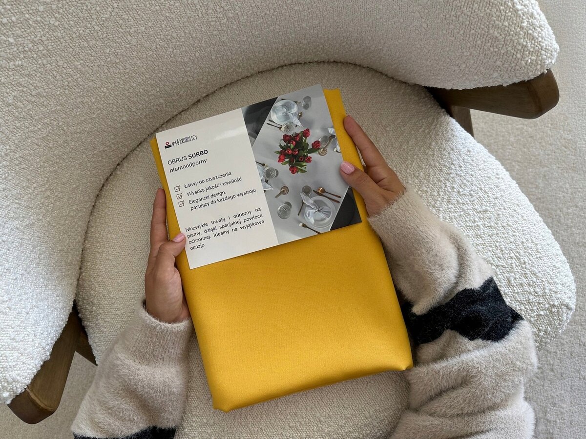 Osoba trzyma musztardowy obrus z etykietą zawierającą tekst: \'OBRUS SURBO plamoodporny\', oraz cechy produktu: \'Łatwy do czyszczenia\', \'Wysoka jakość tkanin\', \'Elegancki design\', \'Pasuje do każdego wnętrza\'.
