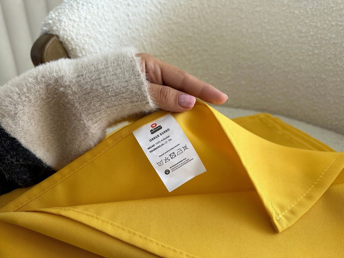 Osoba trzyma musztardowy obrus z widoczną metką. Na metce napisano: \'OBRUS SURBO\', \'120x240 cm\', \'100% POLIESTER\', oraz symbole prania.