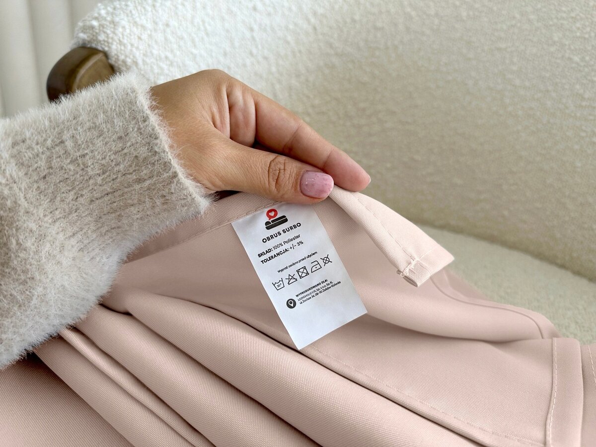 Osoba trzyma różowy materiał z metką, na której widnieje napis: \'OBRUS SURBO, SKŁAD 100% POLIESTER, WYPRODUKOWANO W PL\'.