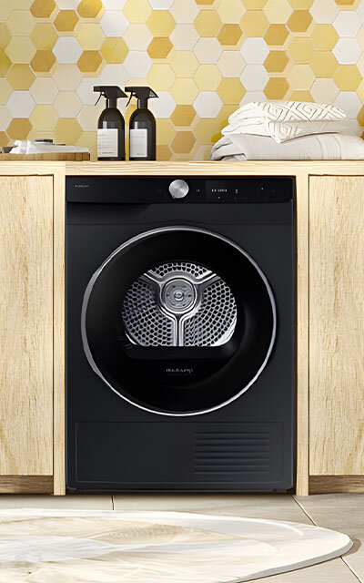 Suszarka DV9XDG6845LBU4 w jasnym pomieszczeniu Ciemna suszarka Samsung w nowoczesnej zabudowie meblowej z jasnego drewna, ustawiona w pralni na tle ściany z żółto-białych heksagonalnych płytek.