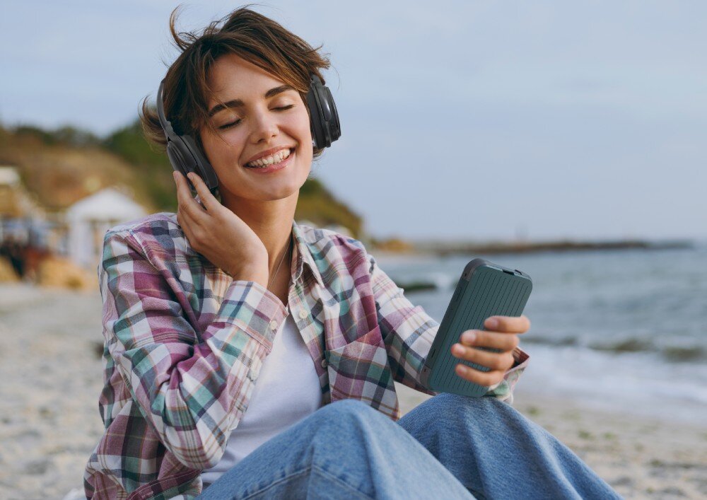 Router ACER Connect M4 5G Uśmiechnięta kobieta w słuchawkach siedząca na plaży i trzymająca w dłoni zielone urządzenie mobilne