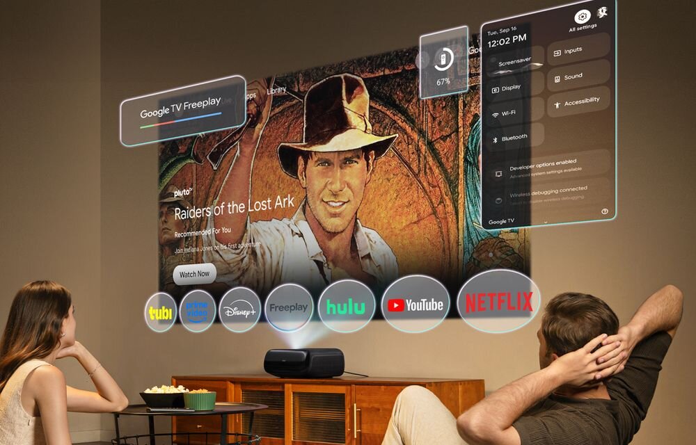 Dwie osoby siedzą przed dużym ekranem, na którym wyświetlany jest interfejs z filmem \'Raiders of the Lost Ark\' oraz ikonami aplikacji takich jak tubi, Disney+, Freeplay, hulu, YouTube i Netflix. Na ekranie widoczne są również ustawienia systemowe oraz godzina 12:02 PM.