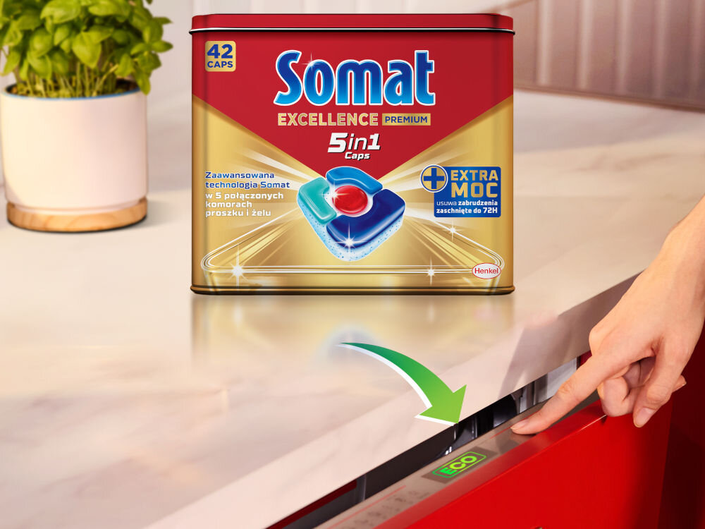 Kapsułki do zmywarki SOMAT Excellence Premium 5w1 - 42 sztuki + Puszka Opakowanie Somat Excellence Premium (42 Caps) na kuchennym blacie, a w tle dłoń włączająca zmywarkę przyciskiem z oznaczeniem ECO, promujące ekologiczne mycie naczyń