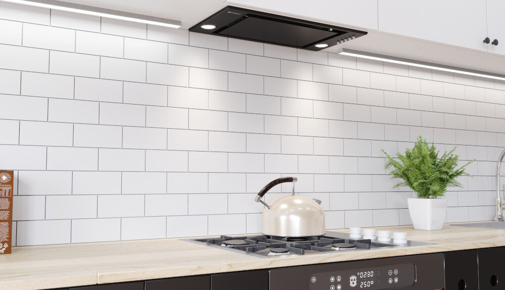 OKAP GLOBALO RETIO 60.2 BLACK Na zdjęciu pokazano kuchnię z białą płytką na ścianie i jasnym blatem, gdzie centralnym punktem jest czajnik stojący na płycie gazowej. Nad kuchenką widoczny jest zintegrowany, czarny okap sufitowy o minimalistycznym designie, dyskretnie wkomponowany w zabudowę meblową. Kuchnia jest bogato oświetlona dzięki długiej, ledowej listwie na tle białych szafek oraz podświetleniu okapu, co tworzy wrażenie czystości i przestrzeni