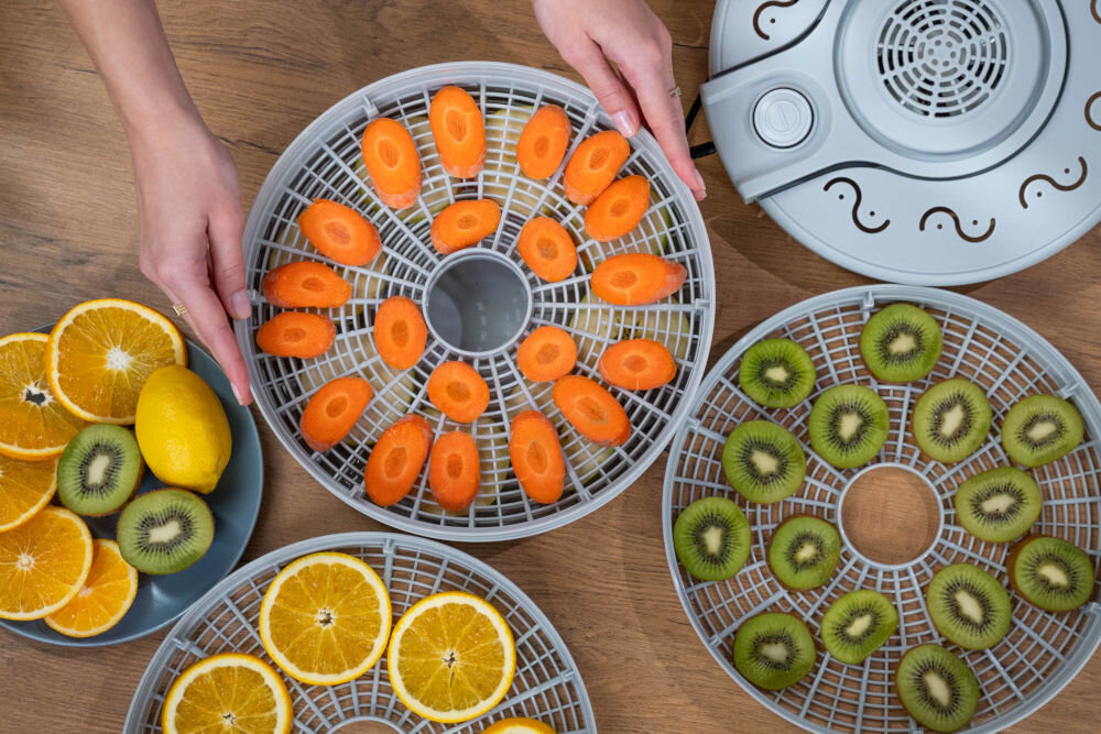 Suszarka do grzybów MILLA HOME FD101E Ujęcie z góry. Dłonie trzymają sito suszarki wypełnione plasterkami marchewki. Obok leżą dwa inne sita: jedno z plastrami kiwi, a drugie z plasterkami pomarańczy i cytryną. Widoczna jest też góra (pokrywa) suszarki.