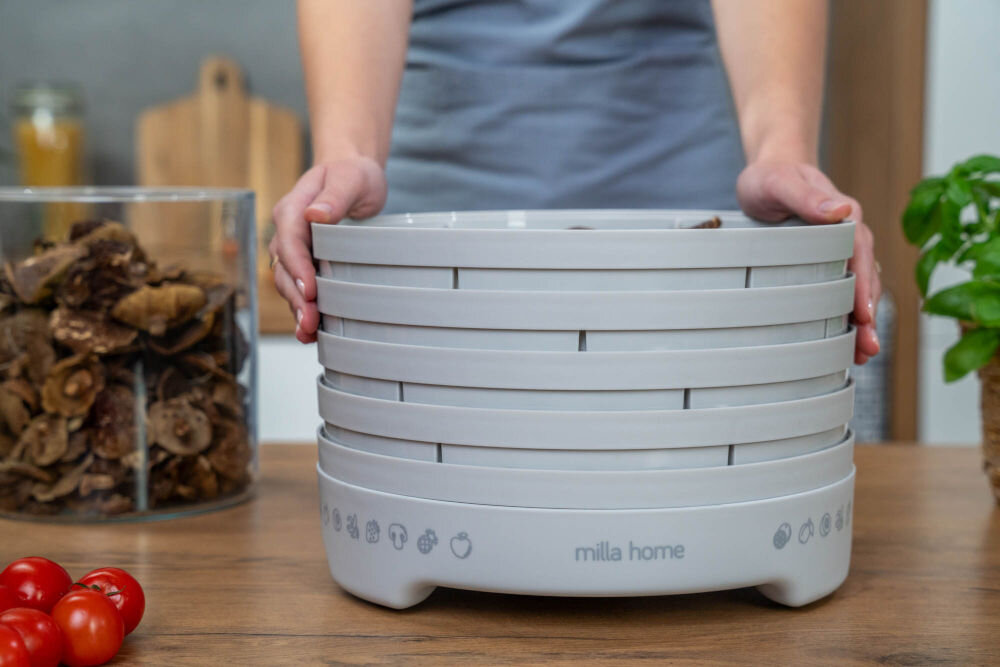Suszarka do grzybów MILLA HOME FD101E Osoba trzyma oburącz szarą suszarkę do żywności Milla Home FD101E z nałożonymi pięcioma sitami. Obok, w dużym szklanym pojemniku, znajdują się ususzone plasterki grzybów.