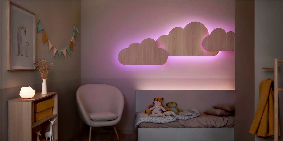 Pokój dziecięcy z łóżkiem, nad którym znajdują się drewniane dekoracje w kształcie chmur podświetlone różowym światłem. Obok łóżka stoi fotel, a na komodzie znajduje się lampa i wazon z suszonymi kwiatami.