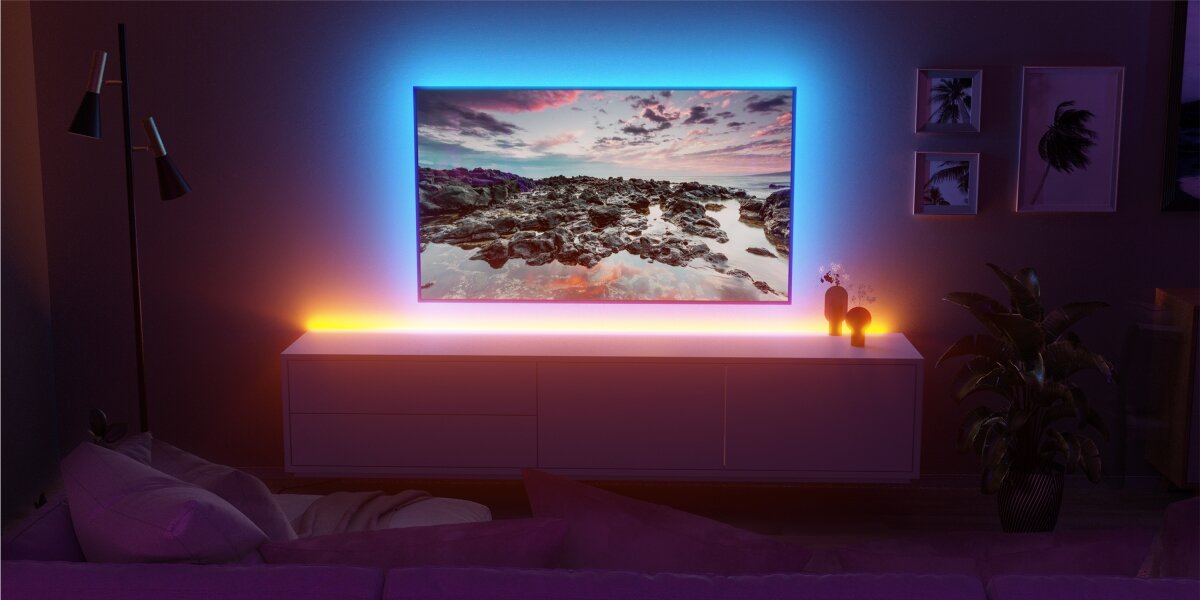 Pokój z telewizorem na ścianie, wokół którego świeci kolorowa taśma LED. Na ekranie wyświetlany jest krajobraz z kamienistym wybrzeżem i kolorowym niebem.