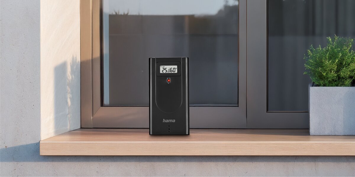 Stacja pogodowa HAMA Usedom Czarny Bezprzewodowy czujnik zewnętrzny stacji pogodowej Hama z małym ekranem LCD umieszczony na drewnianym parapecie okiennym.