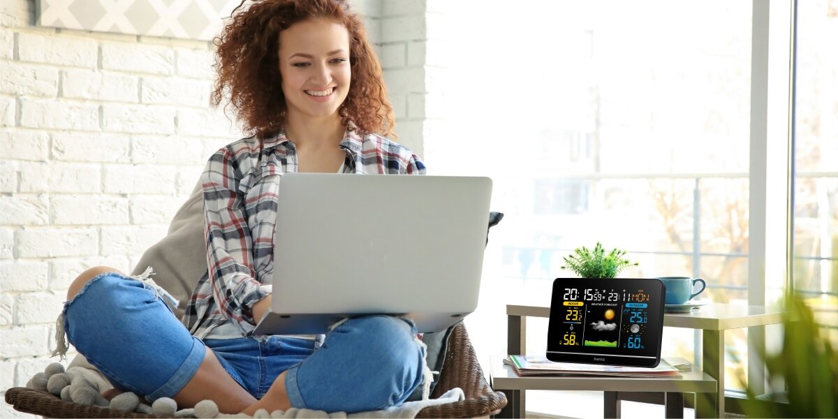 Stacja pogodowa HAMA Usedom Czarny Kobieta pracująca na laptopie w jasnym wnętrzu, obok której na stoliku kawowym stoi stacja pogodowa Hama wskazująca aktualne parametry powietrza.