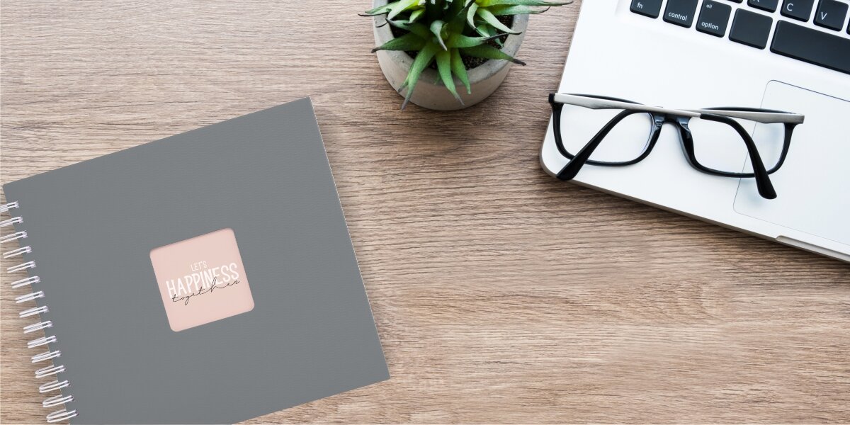 Album HAMA Fine Art Białe kartki Szary (50 stron) Szary album na zdjęcia z napisem „Let’s Happiness Together” leżący na drewnianym biurku obok laptopa, okularów i rośliny doniczkowej.