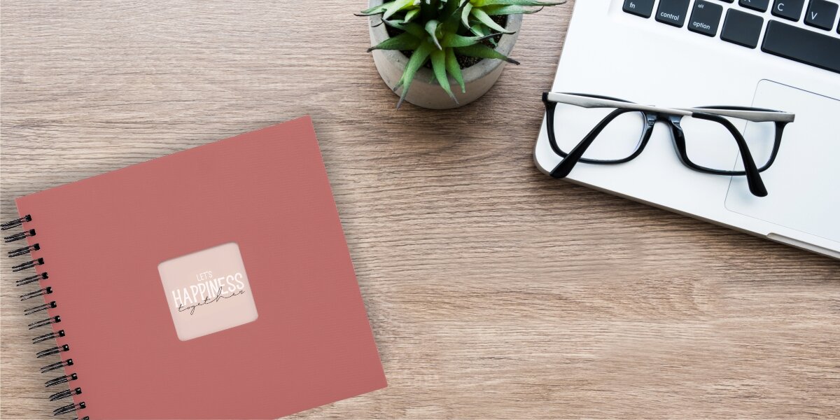 Album HAMA Fine Art Czarne kartki Ceglany (50 stron) Album fotograficzny Let's Happiness Together na drewnianym biurku obok laptopa, okularów i rośliny.