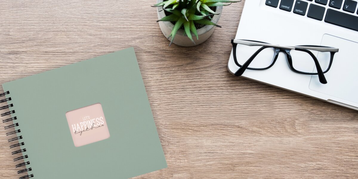 Album HAMA Fine Art Czarne kartki Ceglany (50 stron) Album fotograficzny Let's Happiness Together na drewnianym biurku obok laptopa, okularów i rośliny.