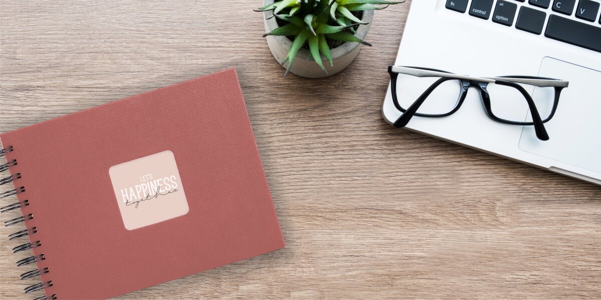 Album HAMA Fine Art Czarne kartki Ceglany (50 stron) Album fotograficzny Let's Happiness Together na drewnianym biurku obok laptopa, okularów i rośliny.