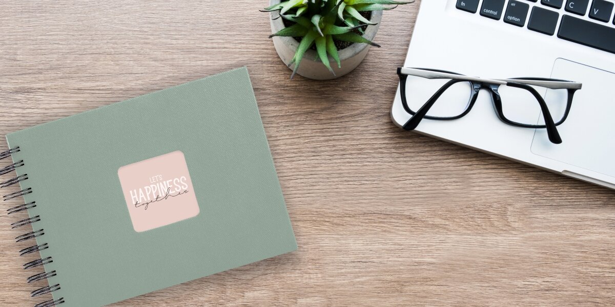 Album HAMA Fine Art 9869 Czarne kartki Szałwiowy (50 stron) Album fotograficzny Let's Happiness Together na drewnianym biurku obok laptopa, okularów i rośliny.