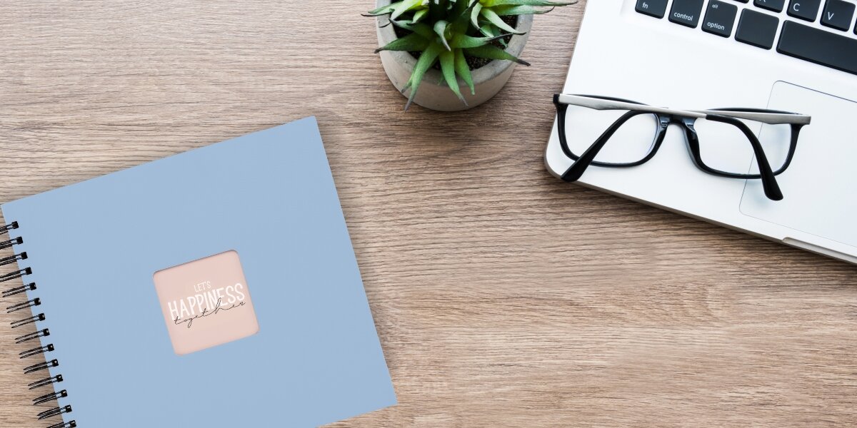 Album HAMA Fine Art Czarne kartki Błękitny (50 stron) Album fotograficzny Let's Happiness Together na drewnianym biurku obok laptopa, okularów i rośliny.