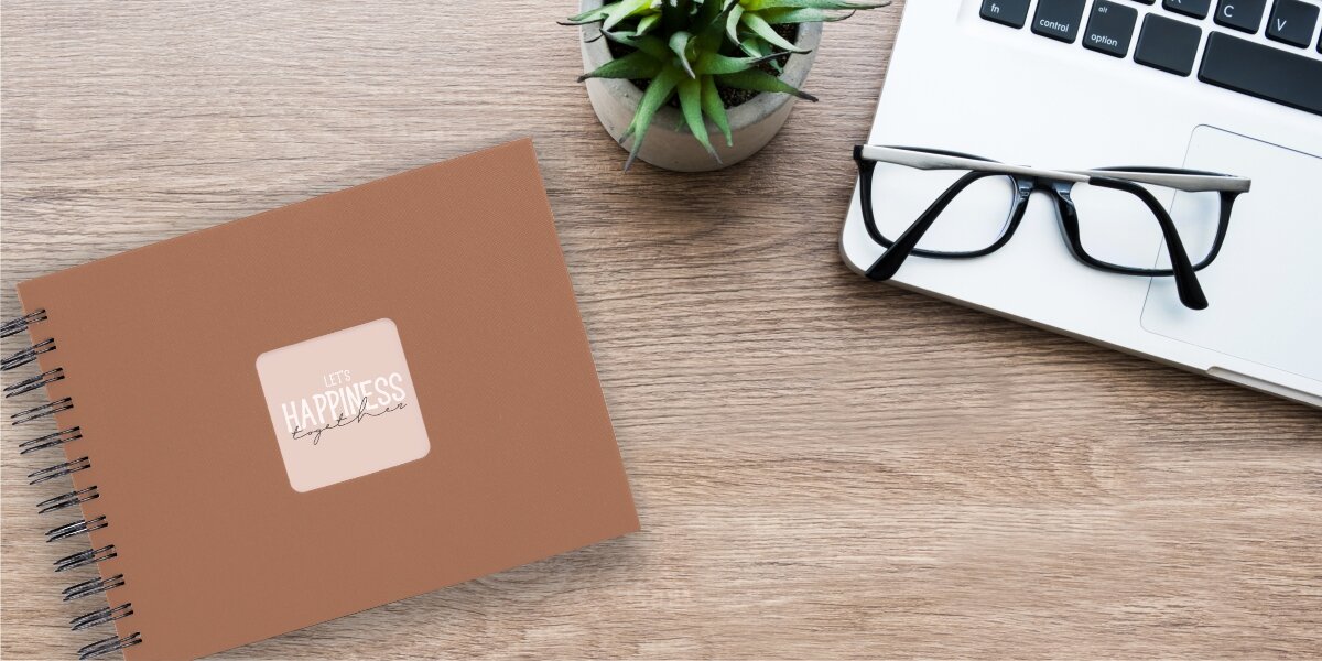 Album HAMA Fine Art Czarne kartki Ceglany (50 stron) Album fotograficzny Let's Happiness Together na drewnianym biurku obok laptopa, okularów i rośliny.