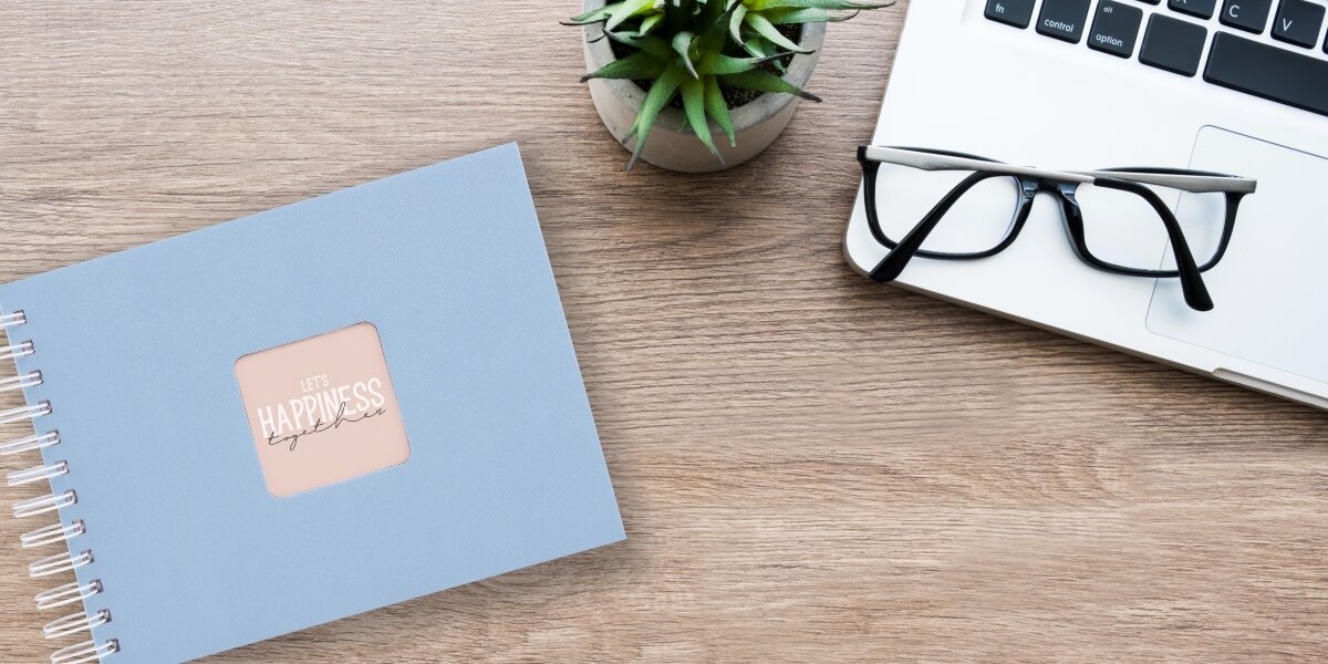 Album HAMA Fine Art Białe kartki Błękitny (50 stron) Album fotograficzny Let's Happiness Together na drewnianym biurku obok laptopa, okularów i rośliny.