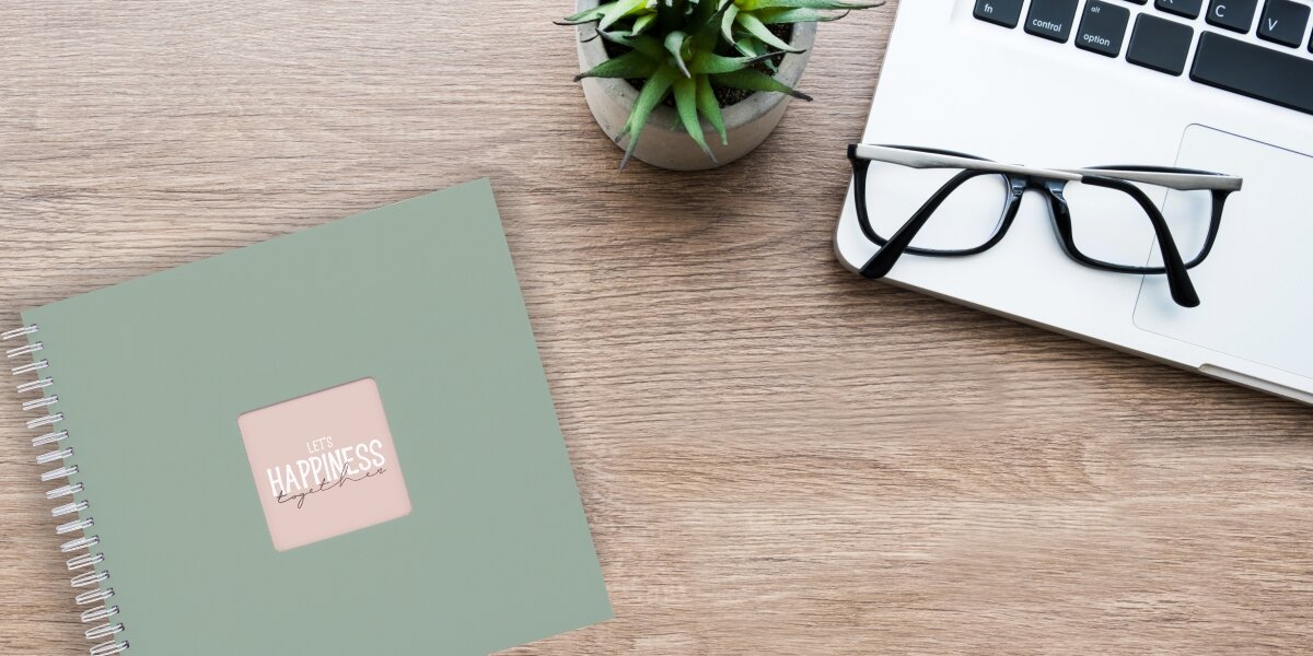 Album HAMA Fine Art 9868 Białe kartki Szarozielony (50 stron) Album fotograficzny Let's Happiness Together na drewnianym biurku obok laptopa, okularów i rośliny.