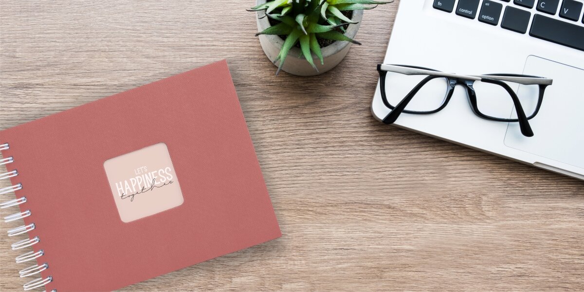 Album HAMA Fine Art Ceglany (50 stron) Album fotograficzny Let's Happiness Together na drewnianym biurku obok laptopa, okularów i rośliny.