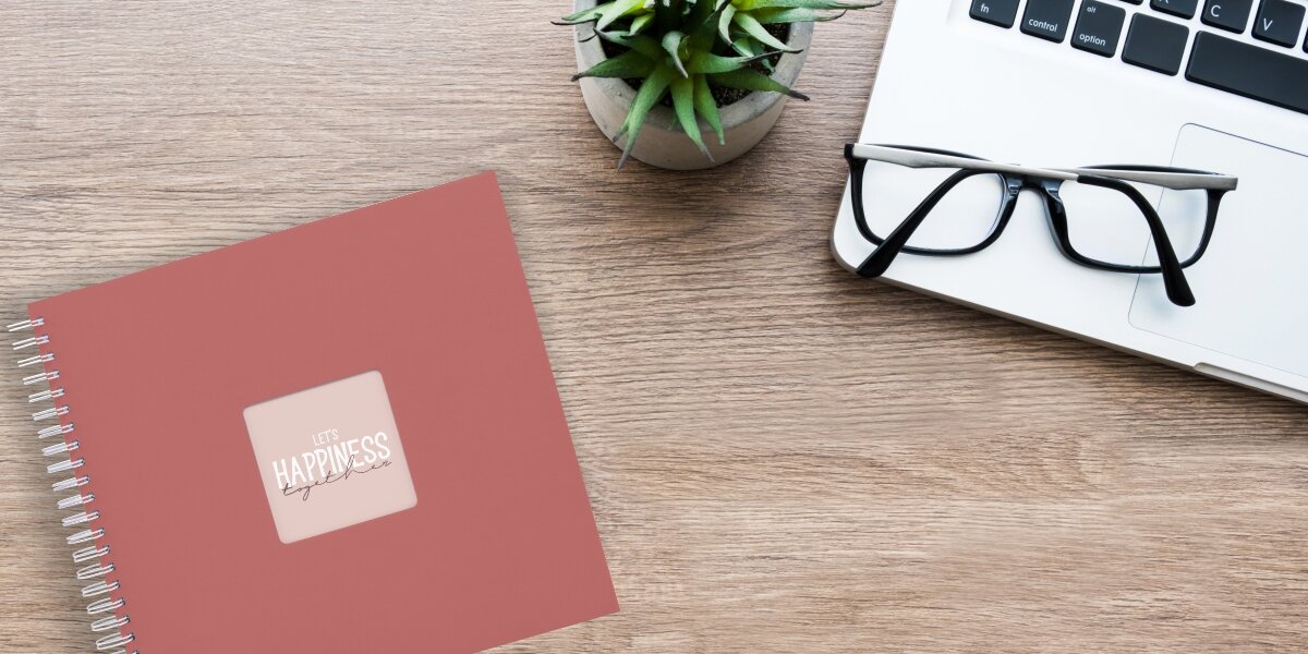 Album HAMA Fine Art Ceglany (50 stron) Album fotograficzny Let's Happiness Together na drewnianym biurku obok laptopa, okularów i rośliny.