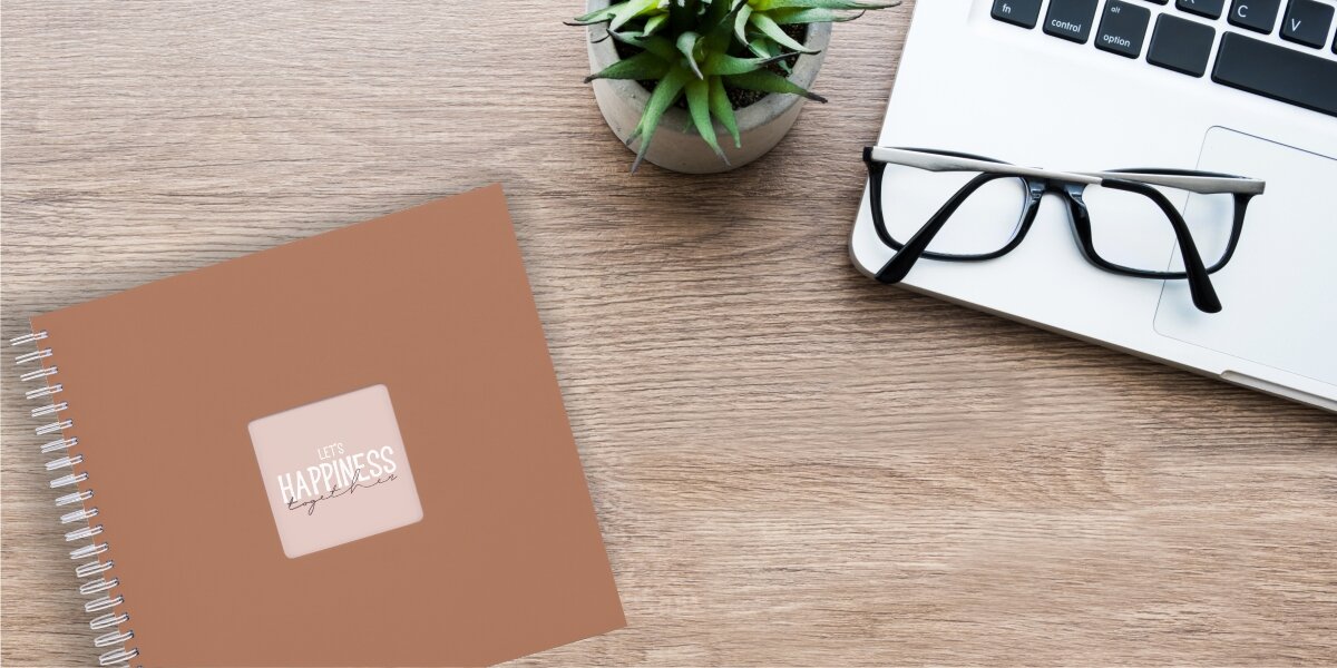 Album HAMA Fine Art Brązowy (50 stron) Album fotograficzny Let's Happiness Together na drewnianym biurku obok laptopa, okularów i rośliny.