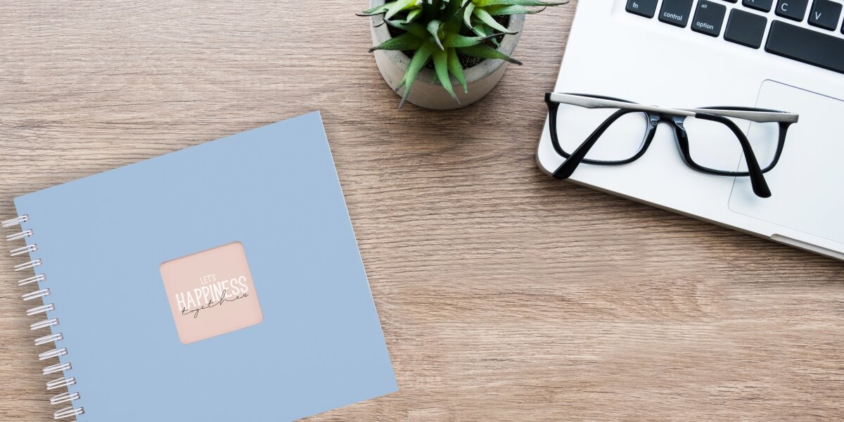 Album HAMA Fine Art Białe kartki Pastelowy błękit (50 stron) Album fotograficzny Let's Happiness Together na drewnianym biurku obok laptopa, okularów i rośliny.