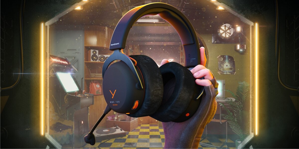 Słuchawki gamingowe beyerdynamic MMX 150 Wireless (czarne z pomarańczowymi akcentami) trzymane w dłoni, z widocznymi dużymi, welurowymi nausznikami i mikrofonem. Tło to stylizowany pokój gracza z automatami do gier i neonami.