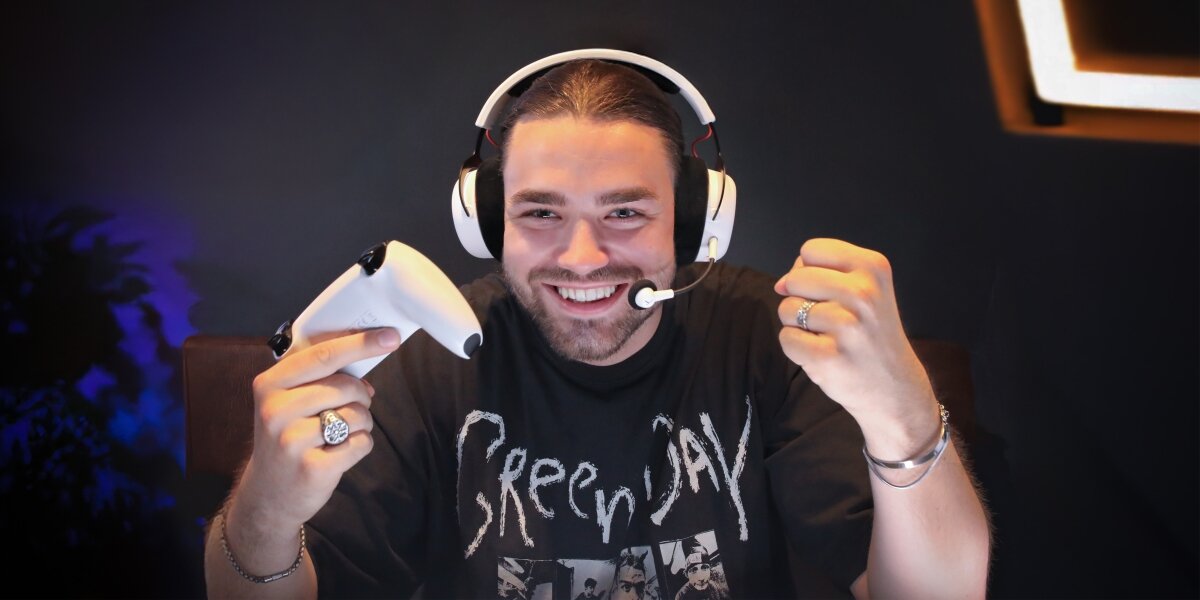 Młody mężczyzna, gracz lub streamer, w białych słuchawkach gamingowych beyerdynamic, trzymający kontroler do konsoli, uśmiecha się i świętuje zwycięstwo. Ubrany w czarną koszulkę Green Day, w tle ciemne, gamingowe otoczenie.