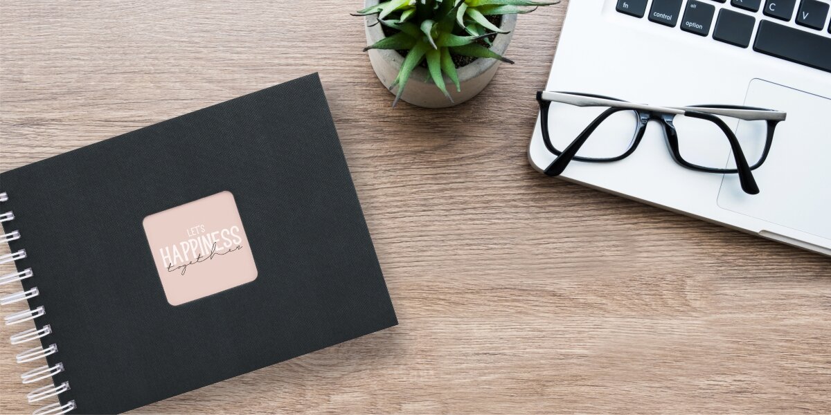 Album HAMA Fine Art 9830 Czarny (50 stron) Album fotograficzny Let's Happiness Together na drewnianym biurku obok laptopa, okularów i rośliny.