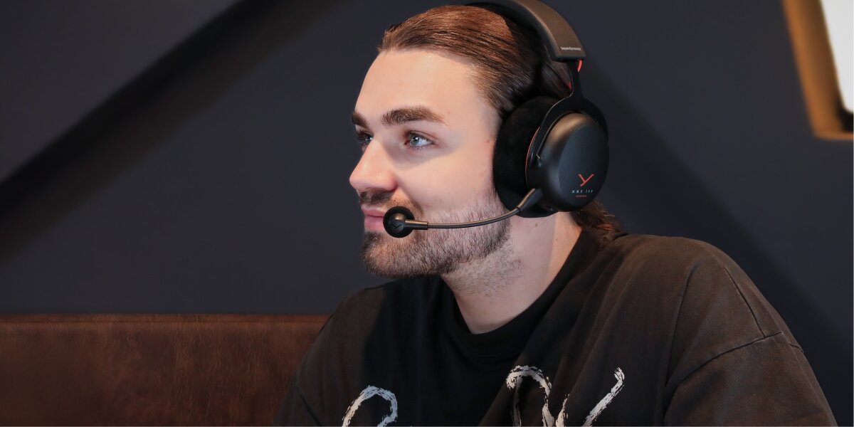 Młody mężczyzna, gracz lub streamer, w białych słuchawkach gamingowych beyerdynamic, trzymający kontroler do konsoli, uśmiecha się i świętuje zwycięstwo. Ubrany w czarną koszulkę Green Day, w tle ciemne, gamingowe otoczenie.