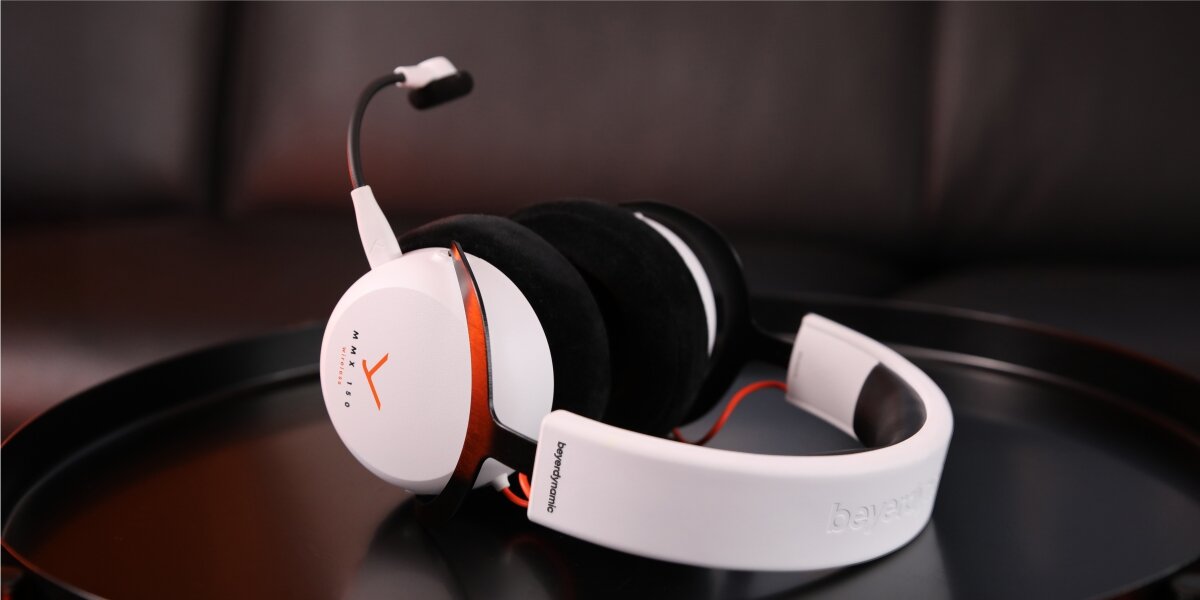 Słuchawki gamingowe beyerdynamic MMX 150 leżące na czarnej, okrągłej podstawce. Podświetlone na czerwono, uwydatniające logo MMX 150 Wireless i odpinany mikrofon na elastycznym pałąku. Scena w klimatycznym, zaciemnionym otoczeniu.