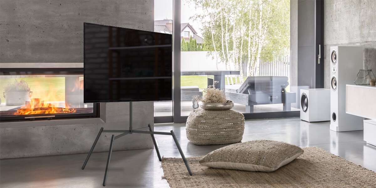 Stojak podłogowy HAMA do TV 60-90 cali 220894 Czarny Nowoczesny stojak pod telewizor typu tripod w industrialnym salonie z kominkiem. Czarny metalowy uchwyt TV na czterech nogach prezentuje się minimalistycznie na tle betonowej ściany i dużych okien.