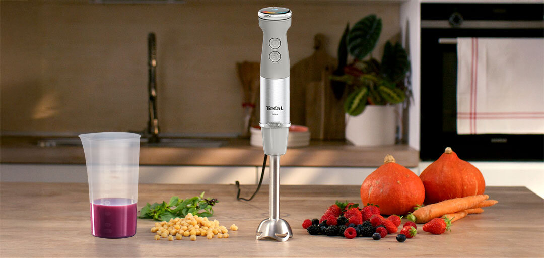 Blender QUICKCHEF+ na kuchennym blacie, obok warzywa i owoce oraz plastikowy kielich z czerwonym płynem.