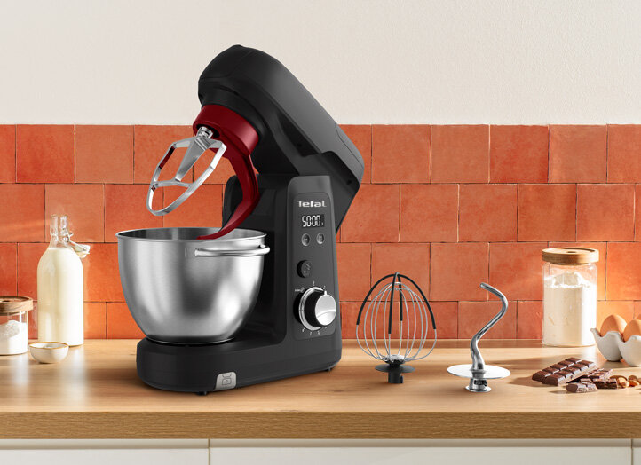 Na blacie kuchennym stoi czarny robot kuchenny z metalową misą i logo Tefal. Obok znajdują się różne akcesoria, takie jak trzepaczka i hak do ciasta, a w tle widać pomarańczowe płytki ścienne.