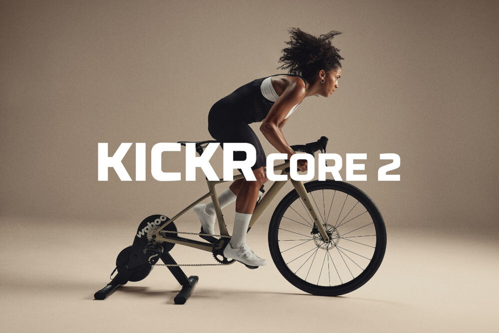 Trenażer rowerowy WAHOO Kickr Core 2 WFBKTR125-3CC jasnobrązowe tło osoba jedzie na rowerze podłączonym do trenażera trening w domu jazda na rowerze wszechstronne zastosowanie