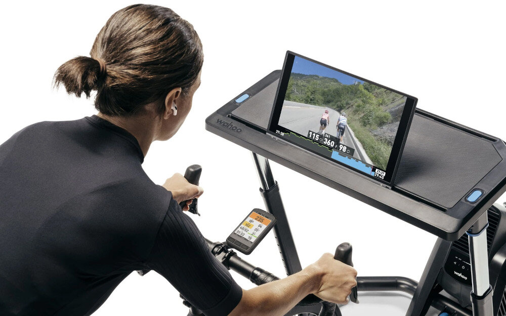 Nawigacja rowerowa WAHOO Elemnt Ace WFCC7 białe tło osoba jedzie na rowerze umieszczonym na trenażerze przed sobą ma stolik z włączonym tabletem na kierownicy umieszczona jest nawigacja wszechstronne zastosowanie planowanie jazdy jazda na rowerze hobby