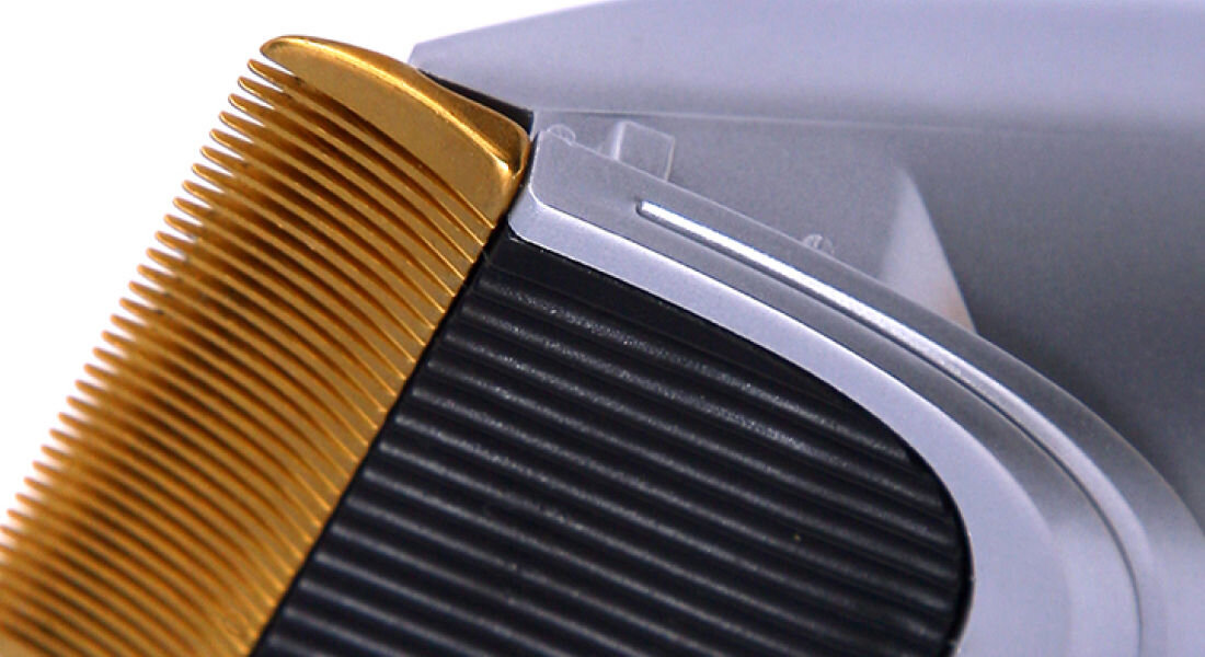 Zbliżenie na ostrze maszynki do strzyżenia z widocznymi złotymi zębami i srebrną obudową. Tło jest rozmyte i szare.