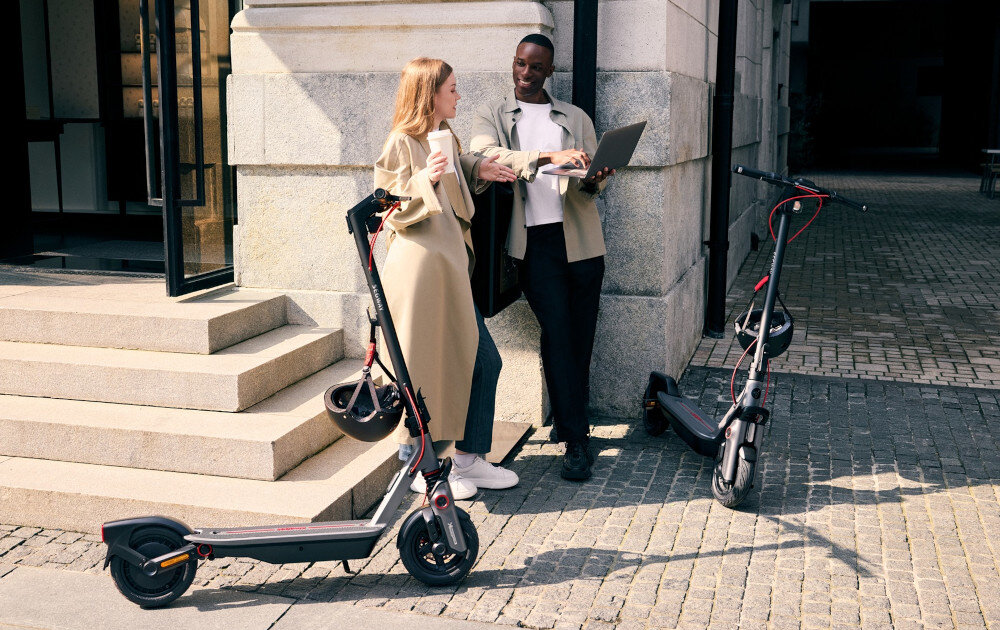 Hulajnoga elektryczna SEGWAY Ninebot F3 Pro D 70km 1200W Amortyzacja 10 cali Czarny miasto budynki chodnik dwie osoby stoją na chodniku obok osób stoją dwie hulajnogi do jazdy po mieście dynamiczne i ekscytujące podróżowanie szereg udogodnień i praktycznych rozwiązań moduł Bluetooth kierunkowskazy składana konstrukcja styl moc wygoda
